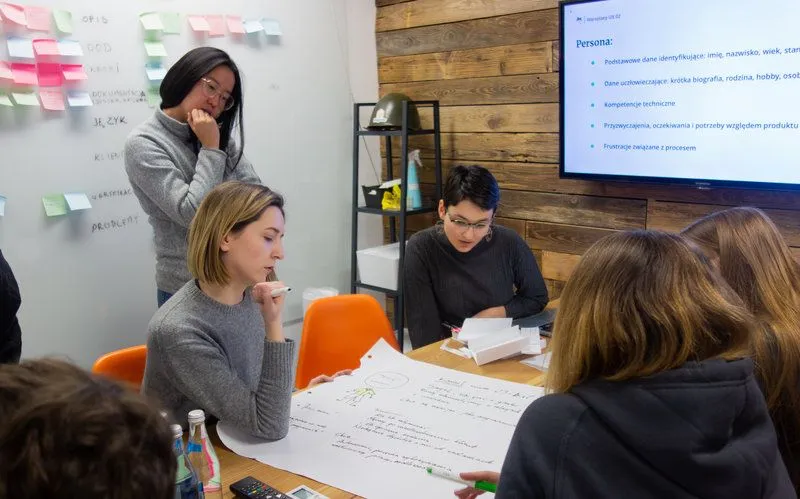 People in conference room, sitting around the table and working together on large piece of paper. A table with post-its and TV with notes displayed on the screen in the background.